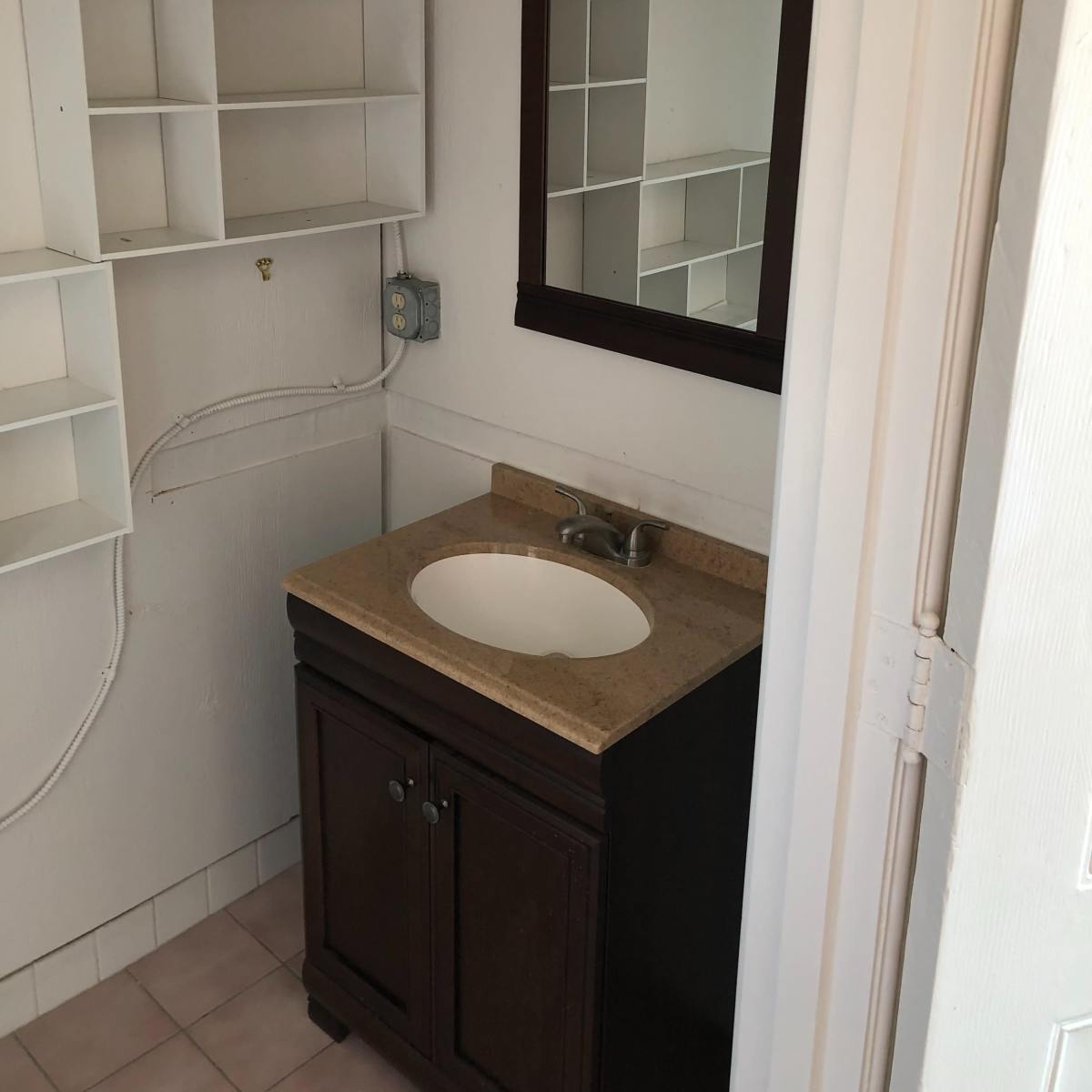 A simple bathroom with a wooden vanity, a sink, a mirror above it, and white shelving on the wall.