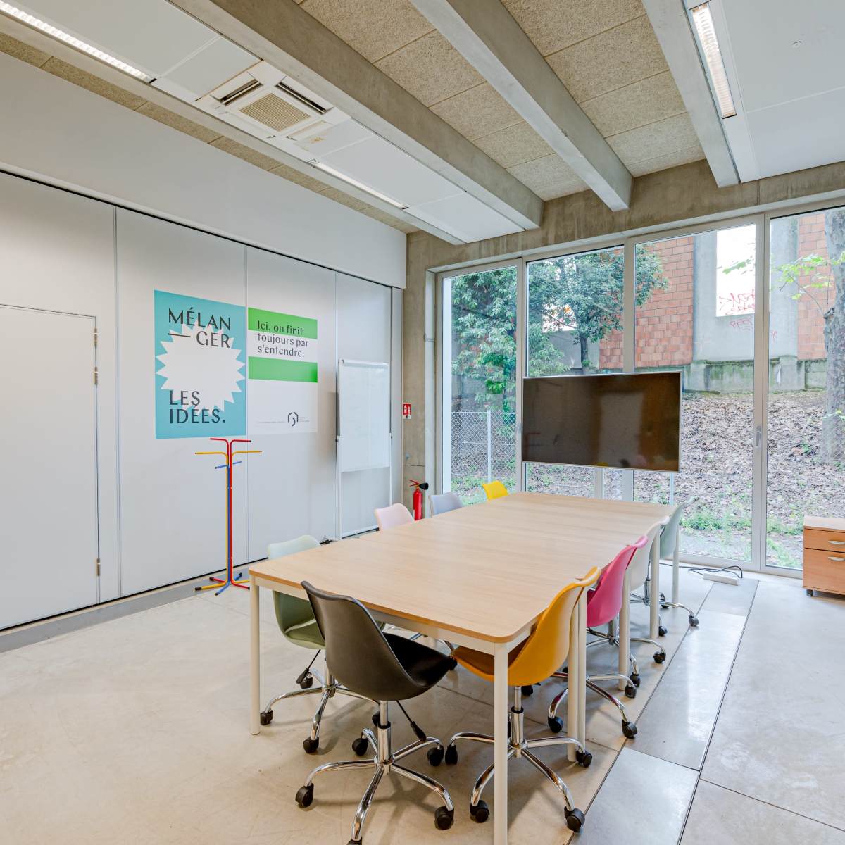 A modern conference room with a large table surrounded by colorful chairs, a wall-mounted screen, and windows overlooking a garden area.