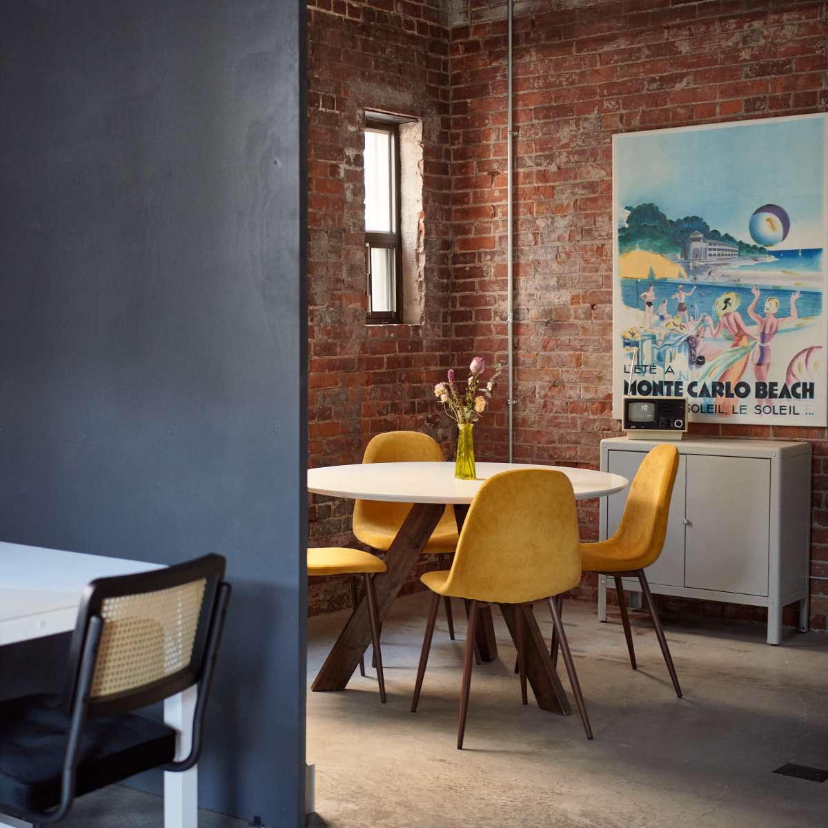 A modern dining area features a white table with black and yellow chairs, surrounded by exposed brick walls and a vintage beach poster.