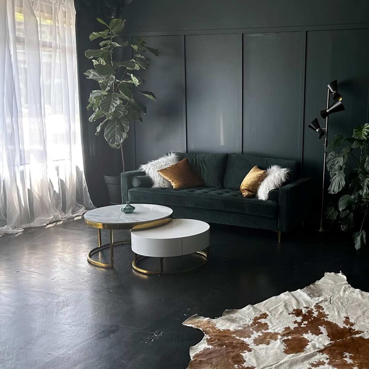 The room features a dark green wall with a green sofa, a round coffee table, a large mirror, a cowhide rug, and various plants.