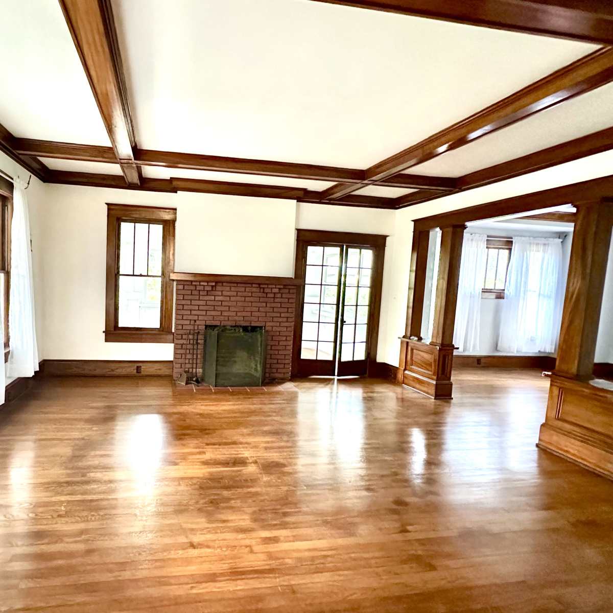 The room features hardwood floors, a brick fireplace, and large windows with sheer white curtains, creating an open and airy space.