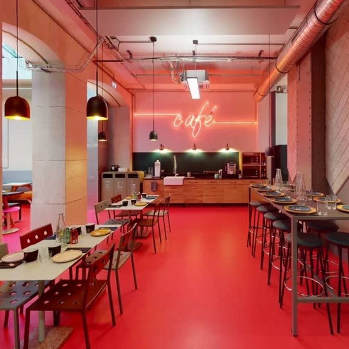 A modern café interior featuring red flooring, wooden furniture, pendant lighting, and a neon sign that reads "café."