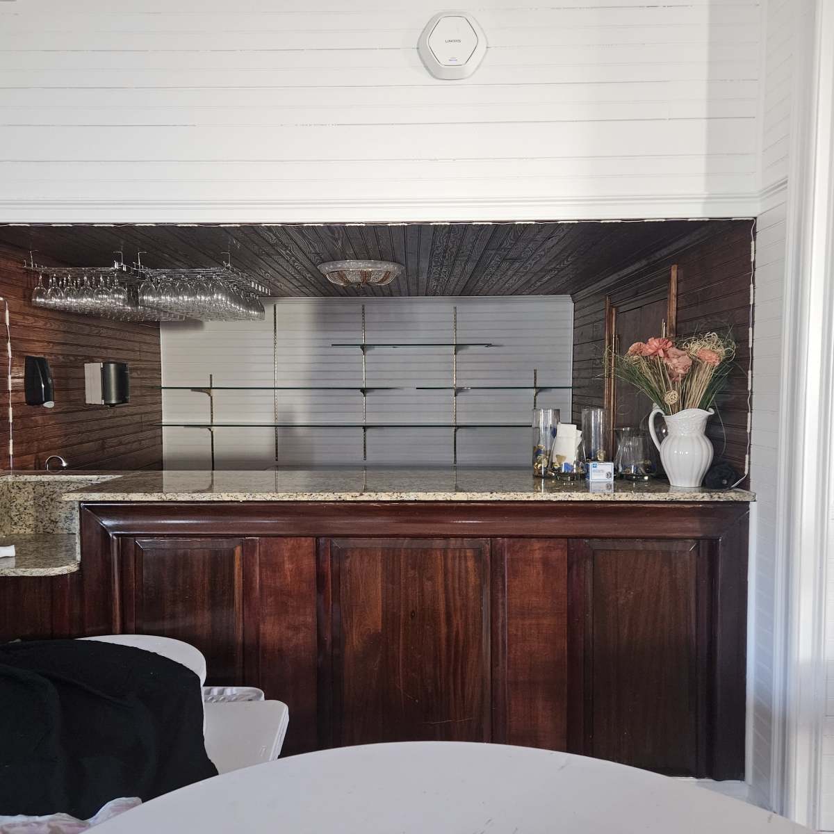 A wooden bar area with a granite countertop, flanked by a set of shelves and an arrangement of glassware and a vase with flowers in the background.