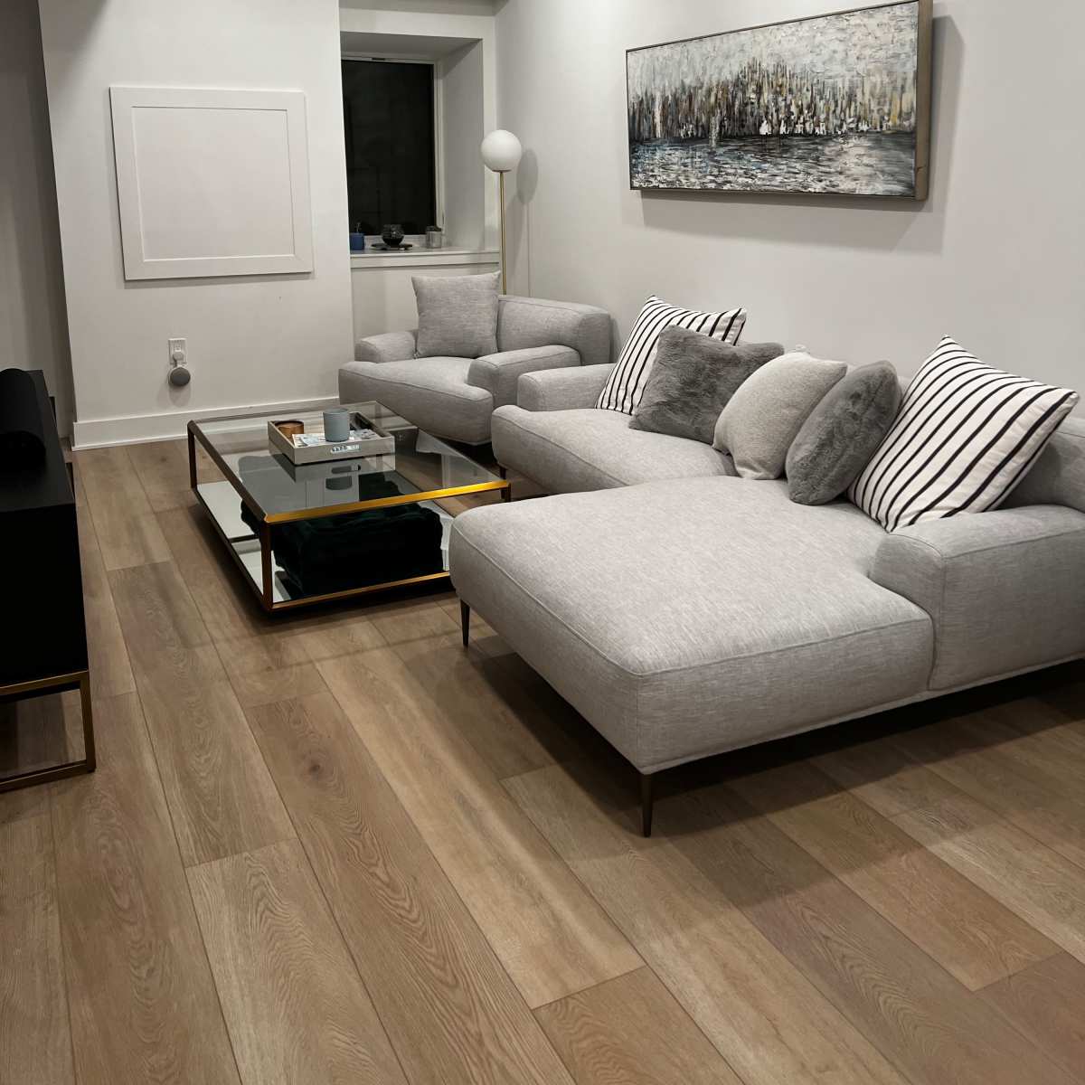 A modern living room featuring two gray sofas, a glass-top coffee table, and a painting above the sofa against a light-colored wall.