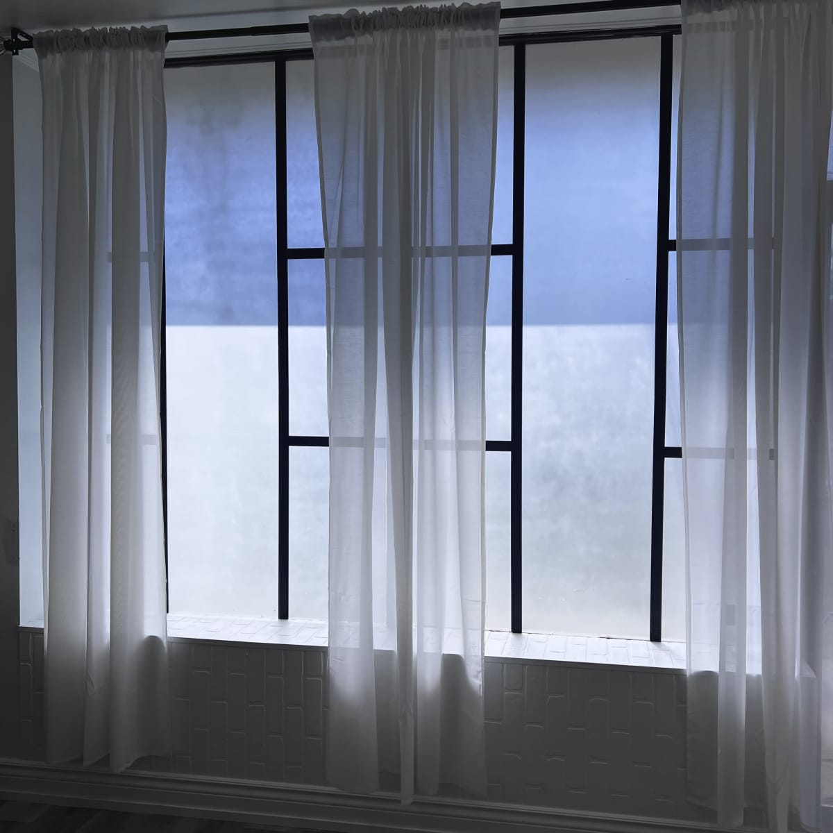 A window with sheer white curtains framing a view of a cloudy sky, bordered by black metal window frames.