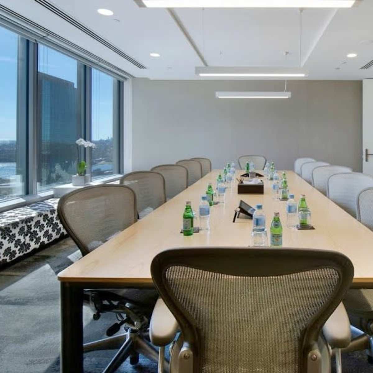 A long conference table with chairs is arranged in a modern office meeting room, featuring large windows that provide a view of the cityscape.