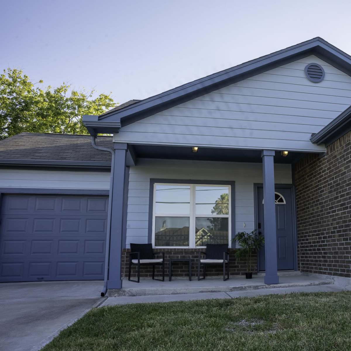 A single-story house with a brick facade and light blue siding, featuring a small porch with two chairs and a garage on the left side.