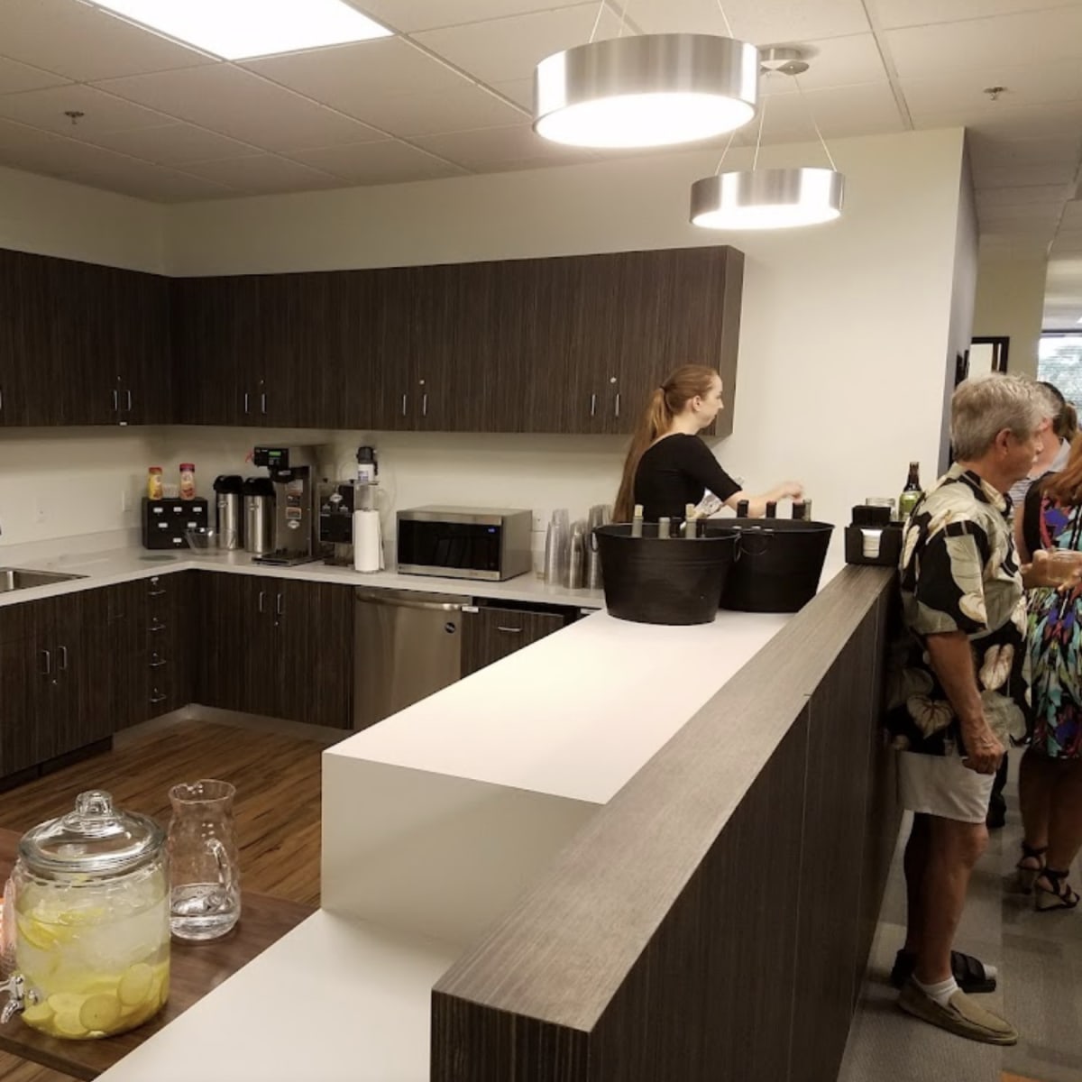 A modern kitchen area features cabinets and appliances with a woman preparing drinks and people socializing nearby.