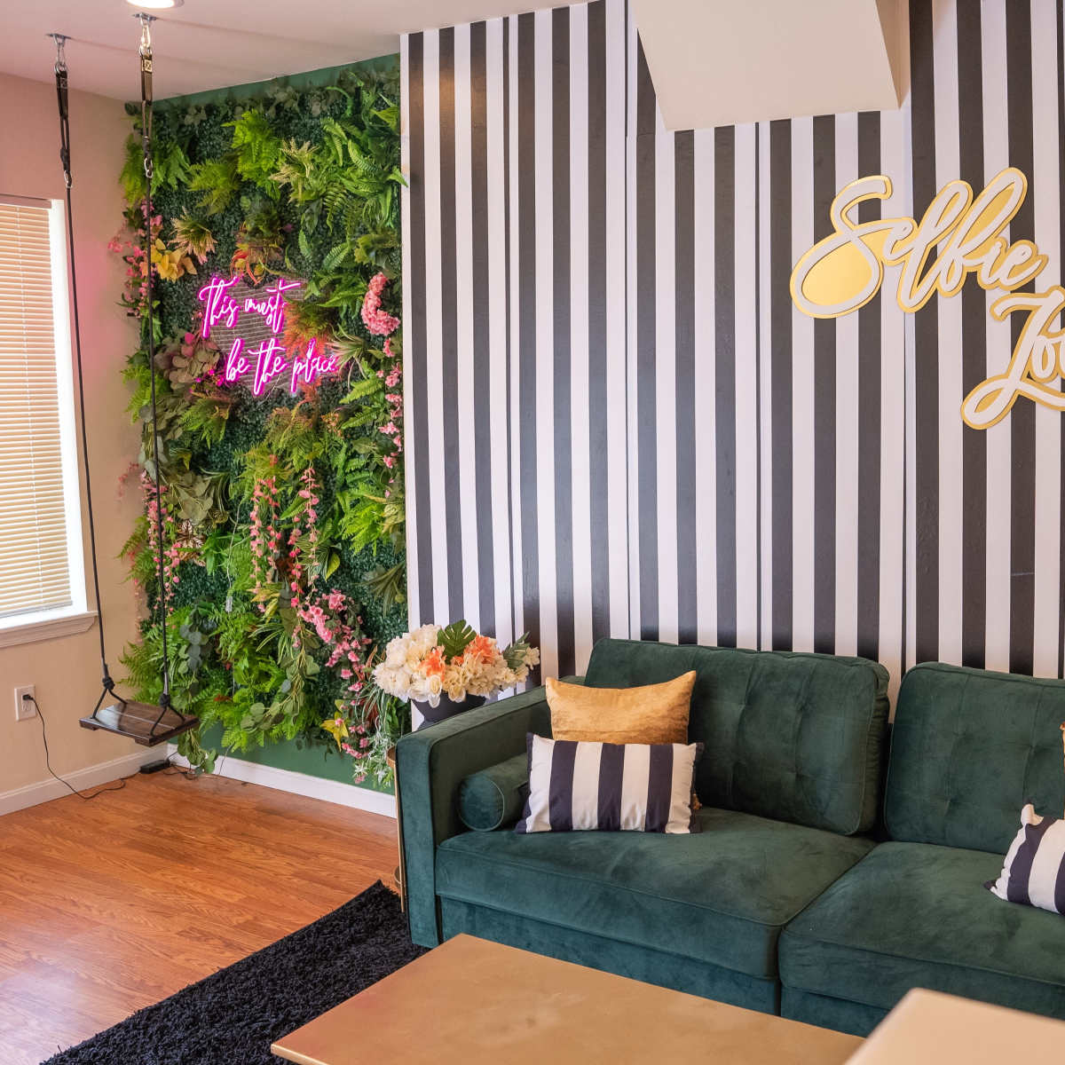 A modern interior with a green velvet sofa, striped wallpaper, and a lively green wall adorned with plants and a neon sign that reads "Selfie Zoo."