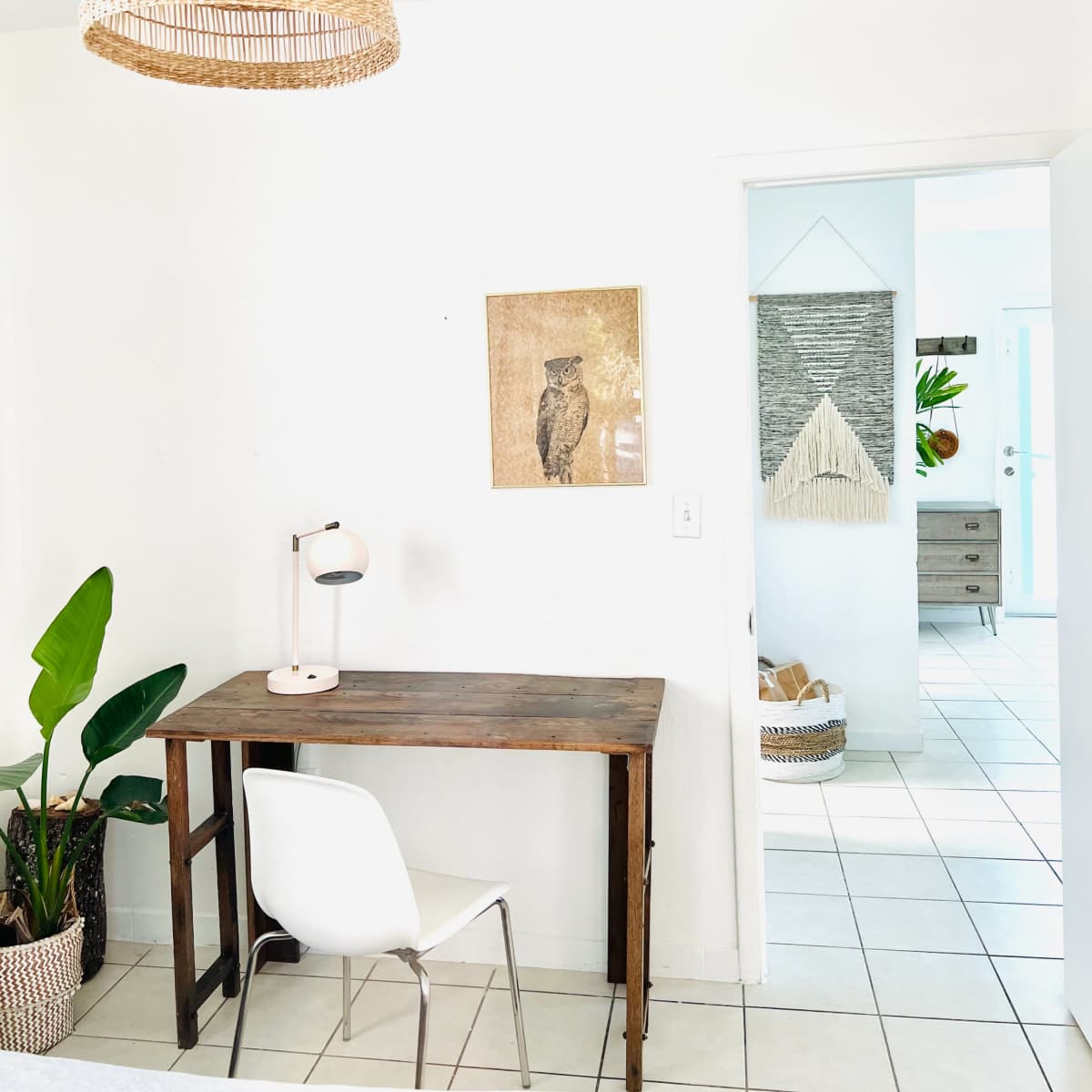 A wooden desk and white chair are positioned against a wall adorned with an owl print, with a large pendant light hanging above in a well-lit room.