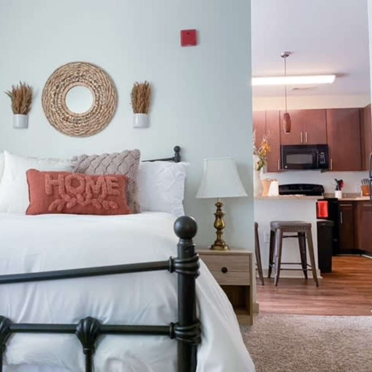 A well-decorated bedroom features a dark metal bed frame with white bedding, adorned with a decorative pillow, and leads to a kitchen area in the background.