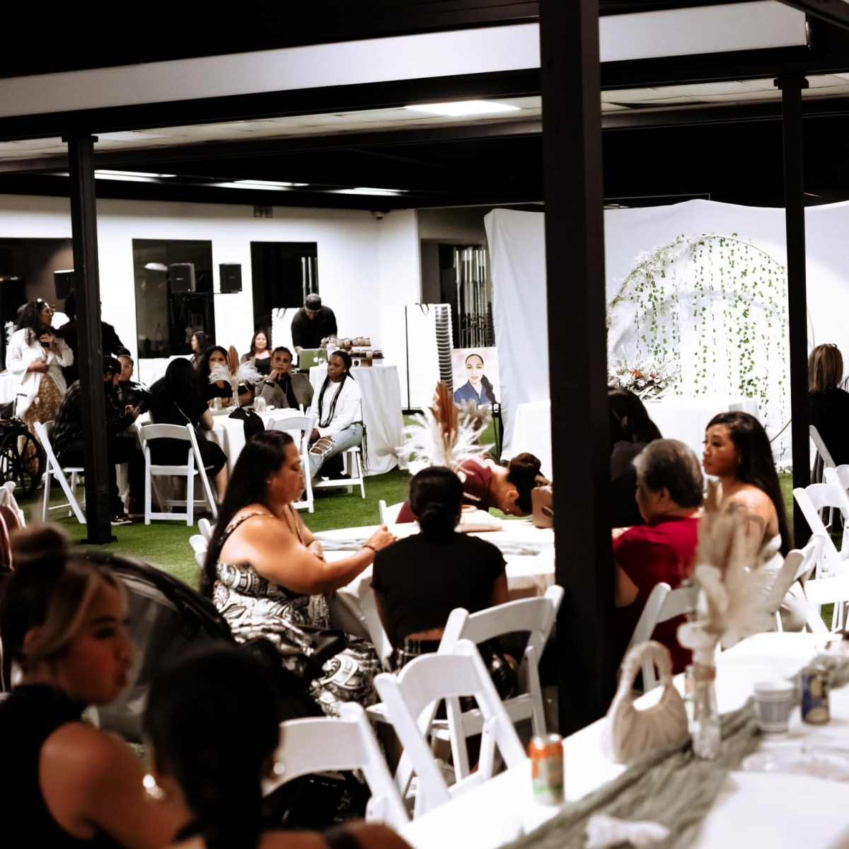 A lively indoor event space with tables arranged for dining and guests engaged in conversation, surrounded by decorations and a backdrop featuring greenery.