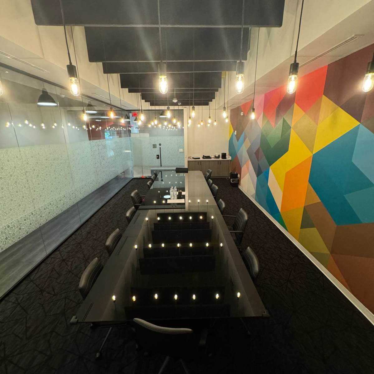 A large, rectangular conference table with a glossy surface and black chairs is positioned in a brightly lit room featuring colorful geometric wall art and suspended light bulbs.