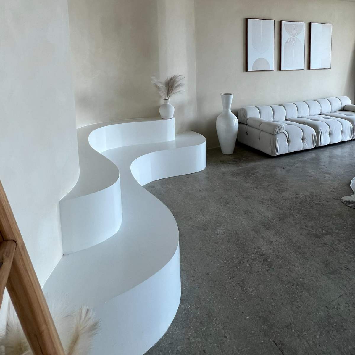 A minimalistic living room featuring a curved white seating area, a white vase, and three framed artworks on the wall.