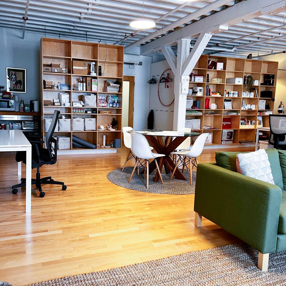A modern open-concept workspace with wooden floors, a large round table surrounded by white chairs, a green couch, and shelves filled with various items.