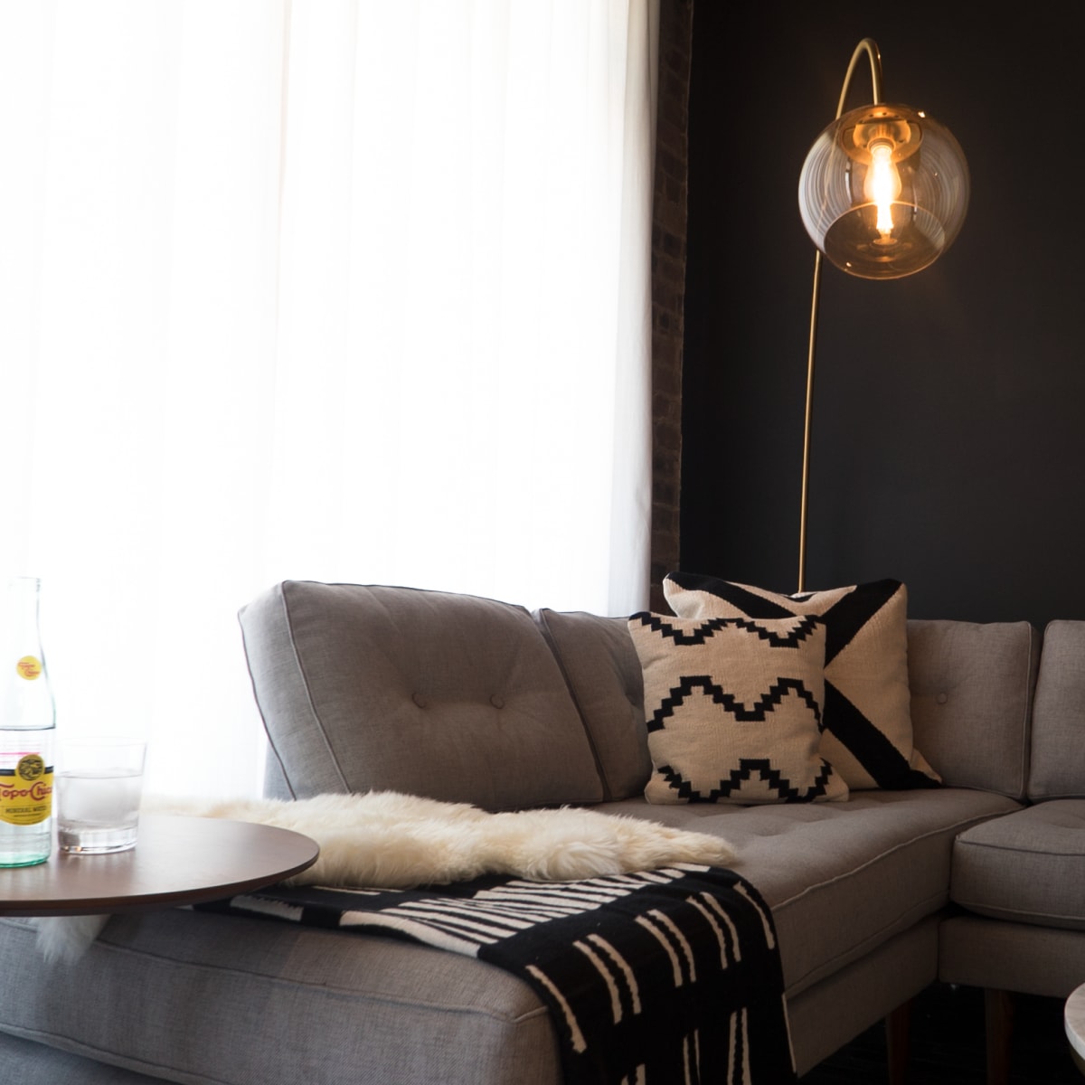 A modern living room features a gray sectional sofa, a round table with drinks, and a floor lamp, all set against a dark wall and illuminated by natural light from large windows.