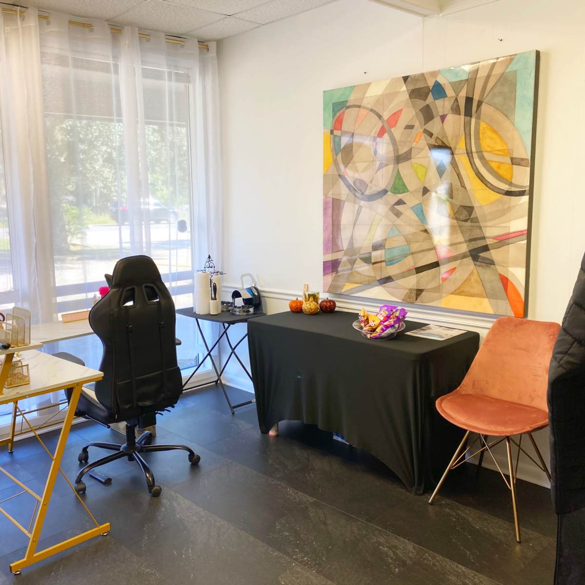 A modern office space features a glass desk and ergonomic chair beside a black table adorned with decorative items, set against a backdrop of large windows and an abstract painting.