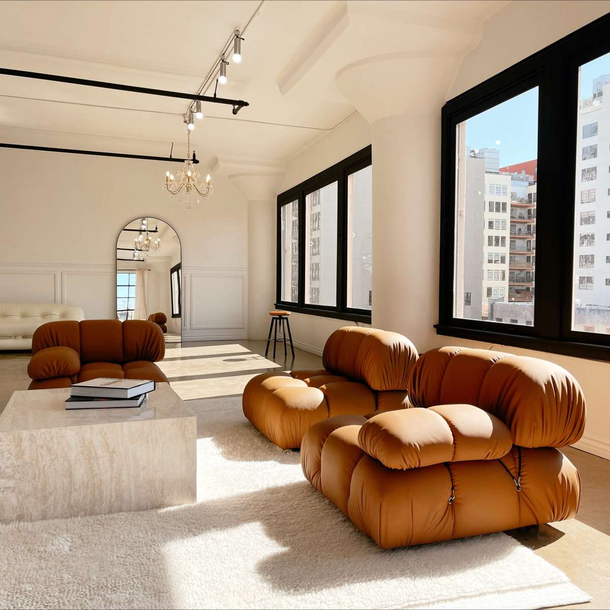 A spacious room with large windows, featuring brown, puffy sofas arranged around a marble coffee table on a light-colored rug.