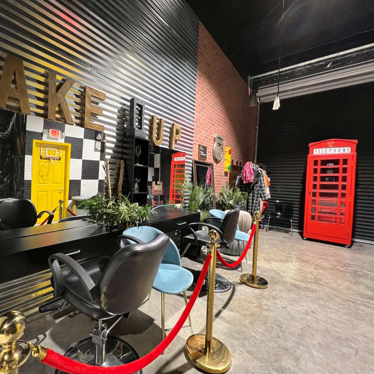 A makeup studio featuring a row of styling chairs, mirrors with large letters spelling "MAKEUP," potted plants, and a red telephone booth in the background.