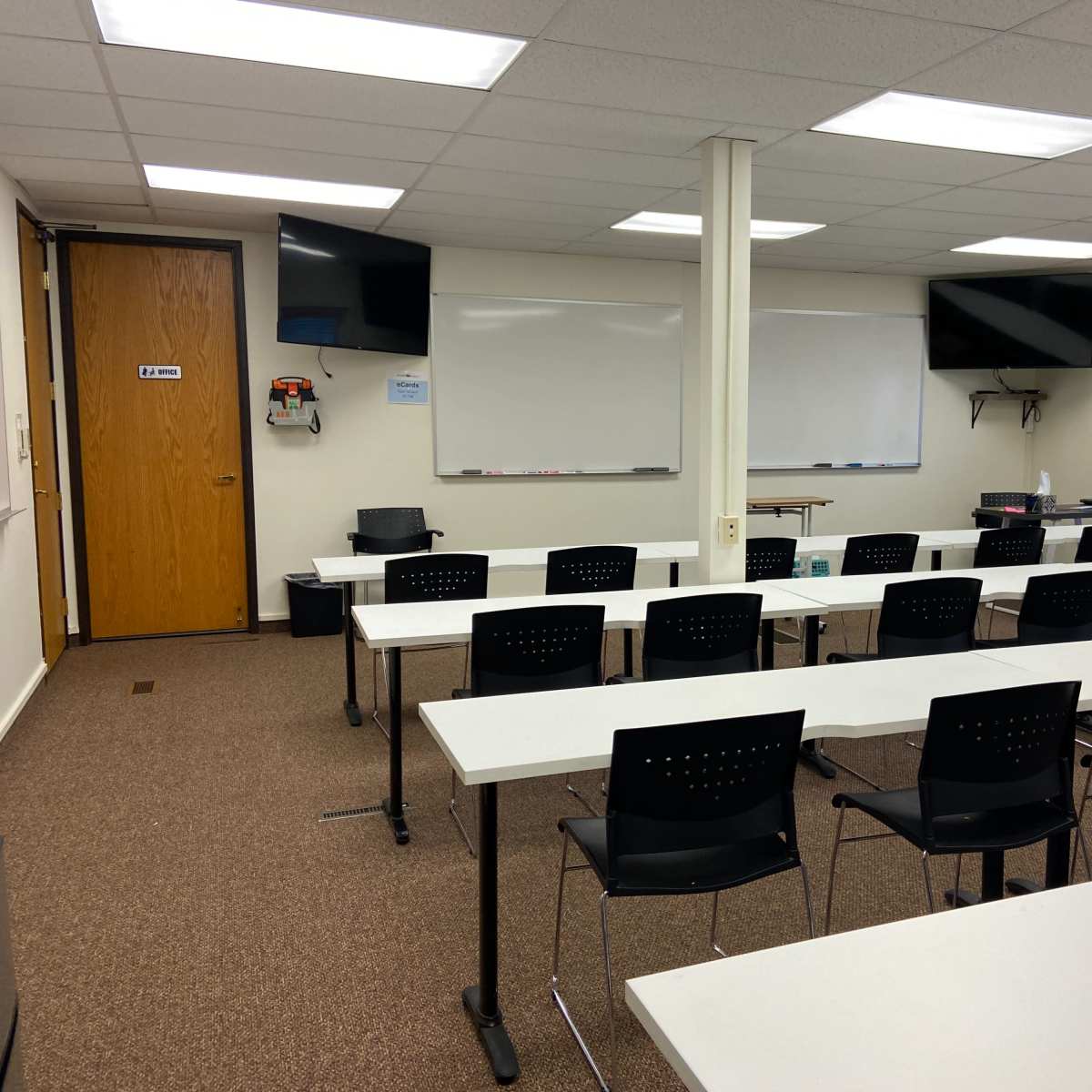 A classroom features several rows of white tables with black chairs, two large whiteboards, and two wall-mounted screens.