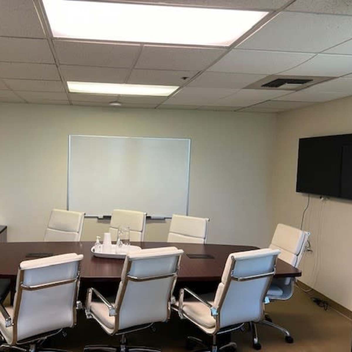 A conference room features a large rectangular table surrounded by eight white ergonomic chairs, a whiteboard on one wall, and a wall-mounted television on another.