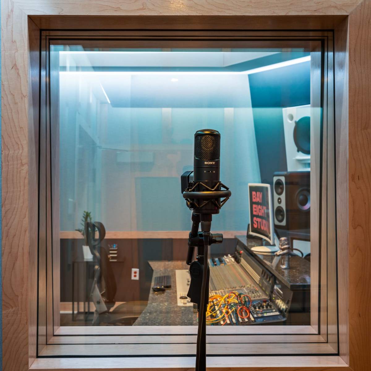 A sound recording studio with a microphone on a stand in front of a glass window, revealing audio equipment and various instruments inside.