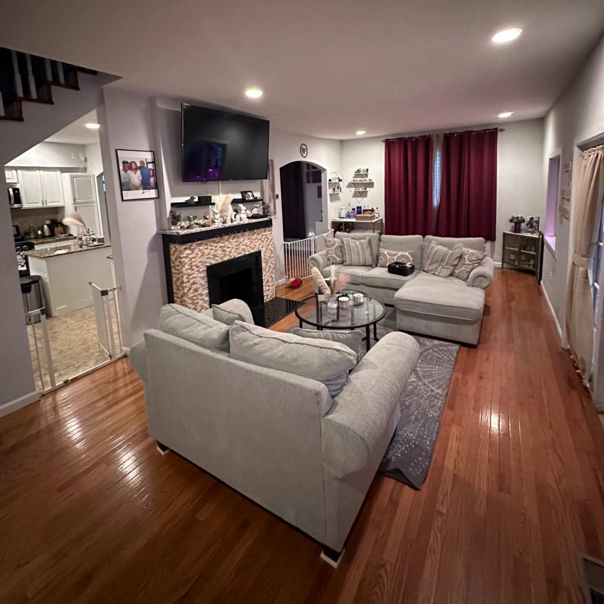 A modern living room with a beige sectional sofa, a coffee table, a TV mounted above a stone fireplace, and red curtains on the windows.