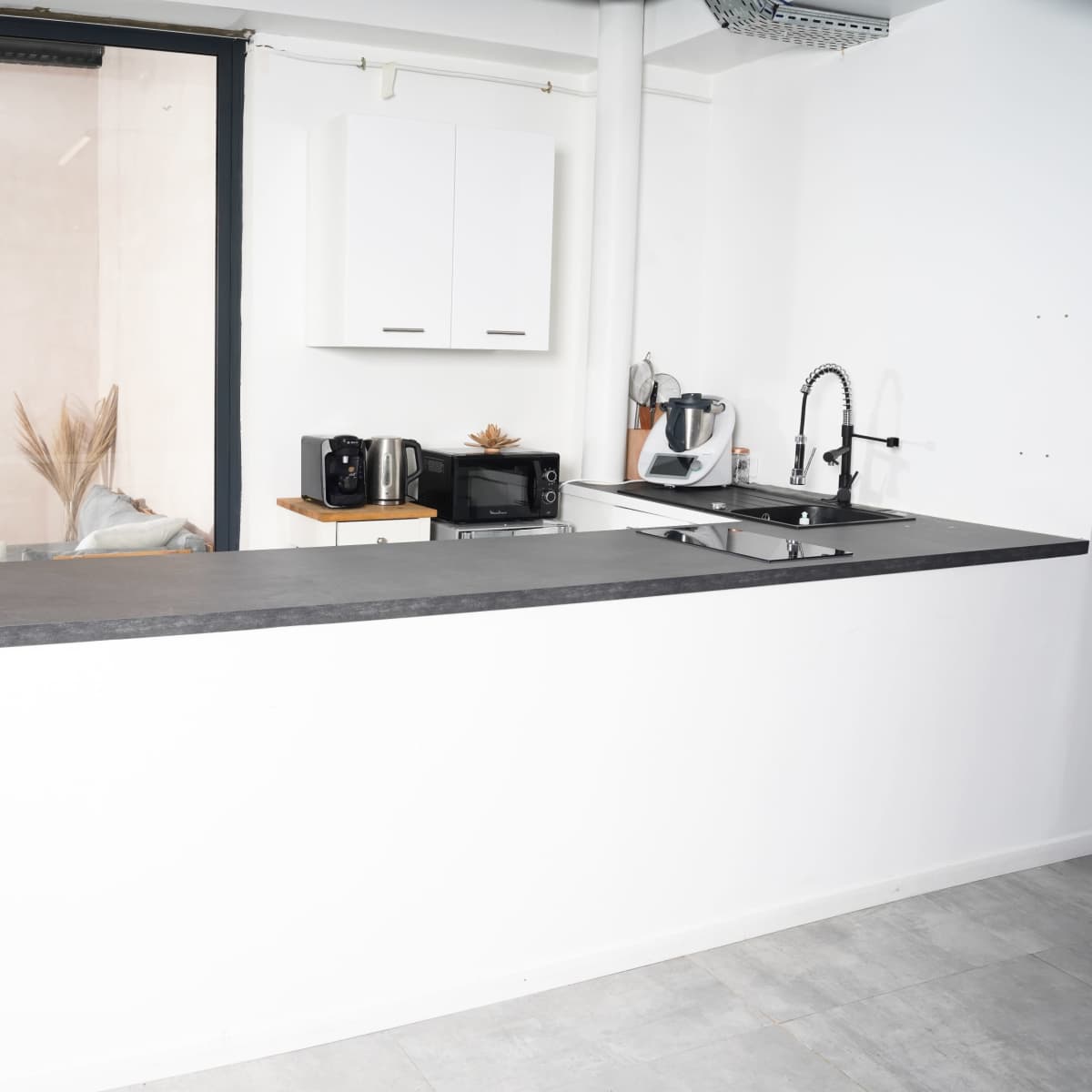 A modern kitchen with a white bar counter, a sink, and small appliances set against a large window displaying plants in the background.