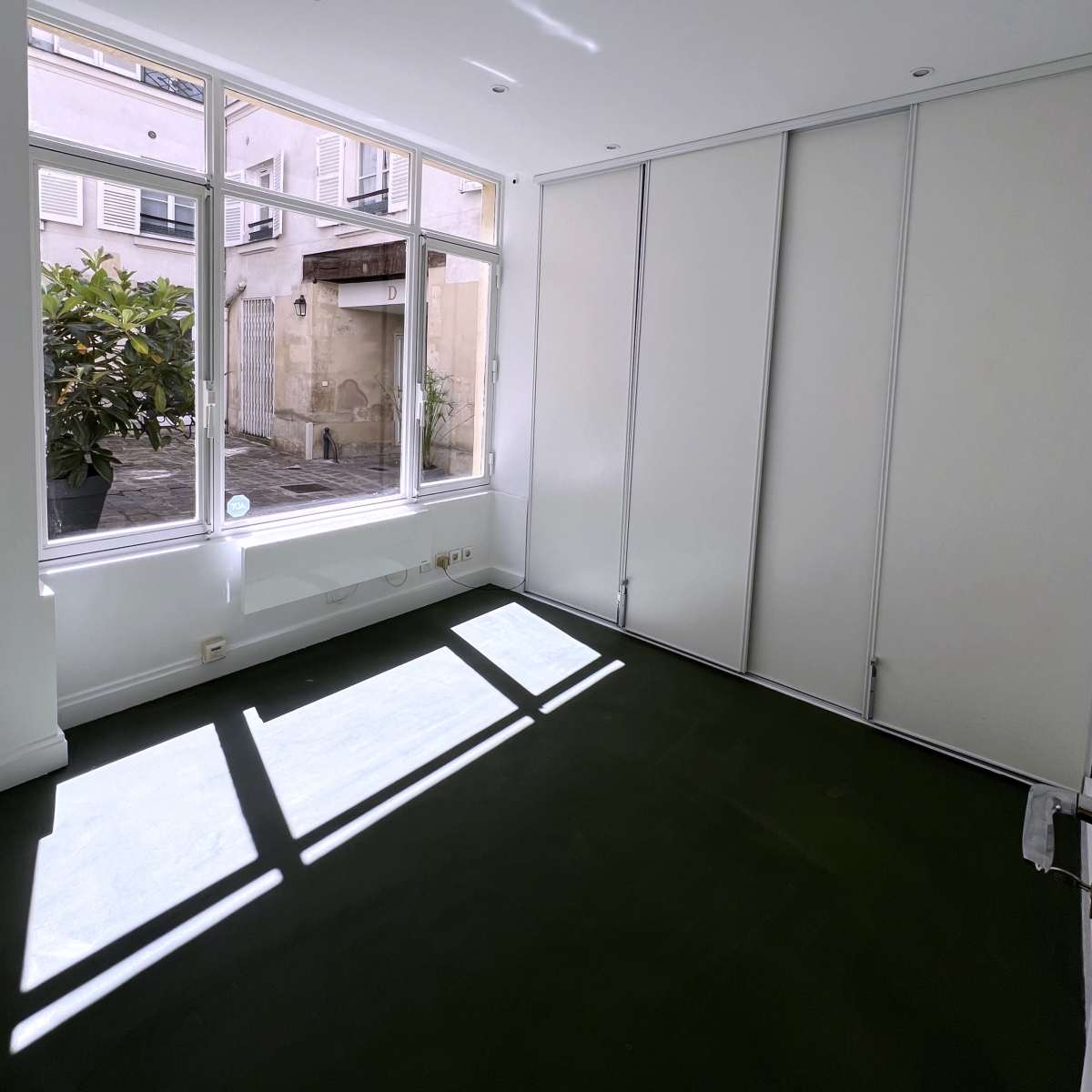 An empty room with large windows allowing natural light to enter, featuring white walls and a dark green floor.