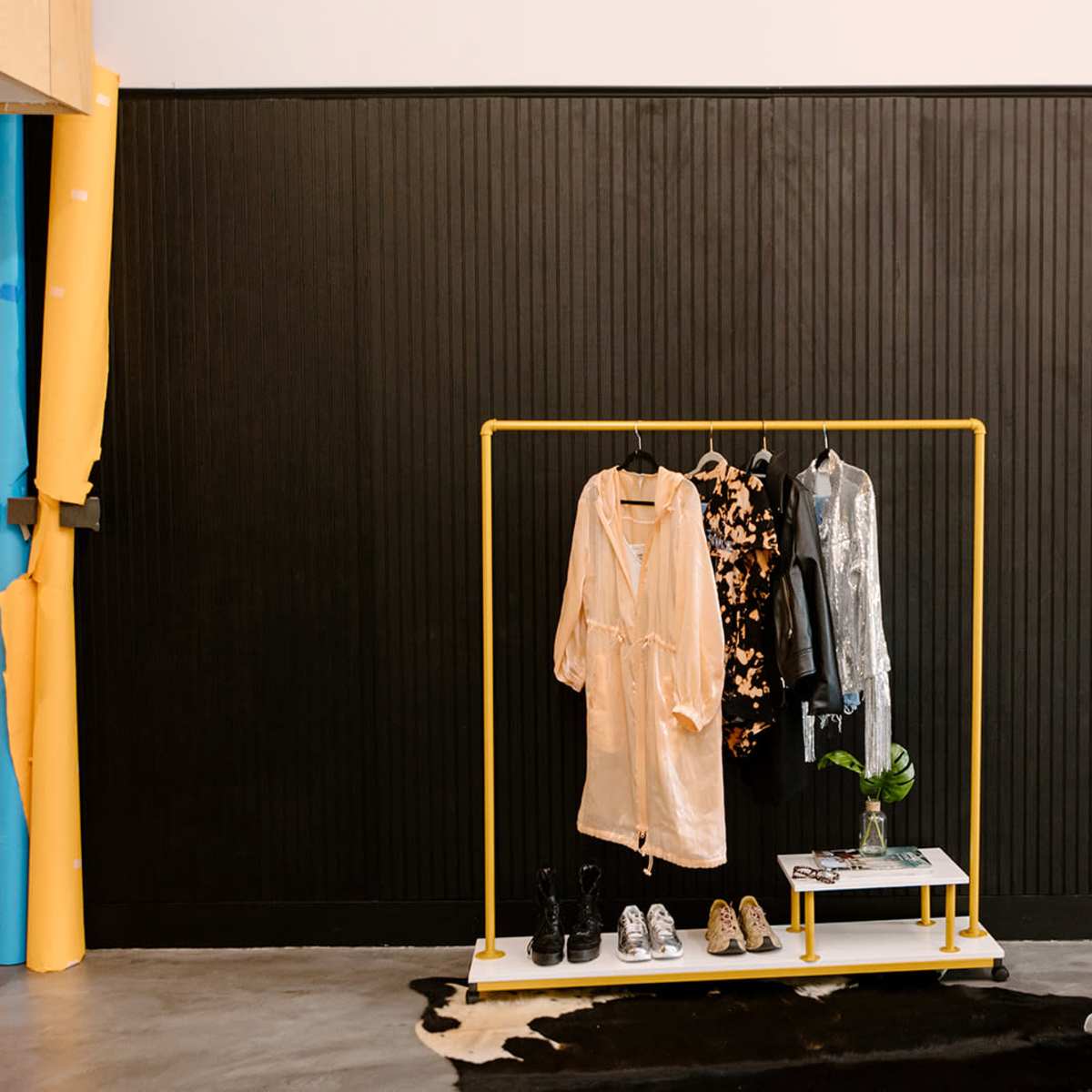 A yellow clothing rack displays three outfits and two pairs of shoes against a black wall in a modern space.