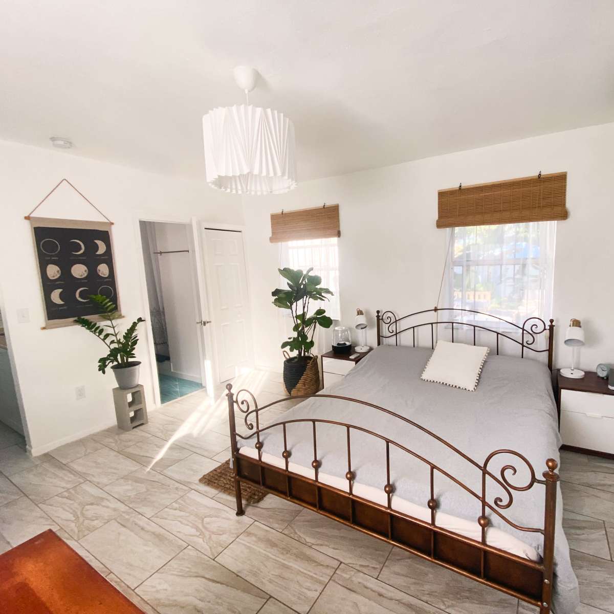 A well-lit bedroom featuring a metal bed frame with a light gray quilt, wooden nightstands, and indoor plants, alongside an open door leading to a bathroom area.