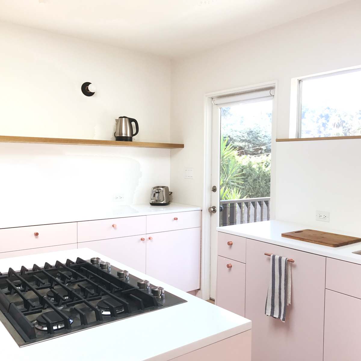 A modern kitchen with pink cabinetry, a gas stovetop, a wooden cutting board, and a large window that opens to an outdoor view.