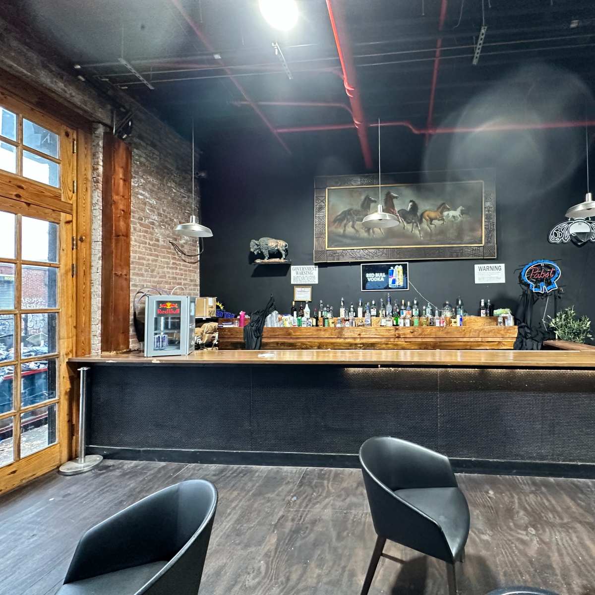 A bar area with a wooden counter, shelves filled with liquor bottles, and two pendant lights hanging above.