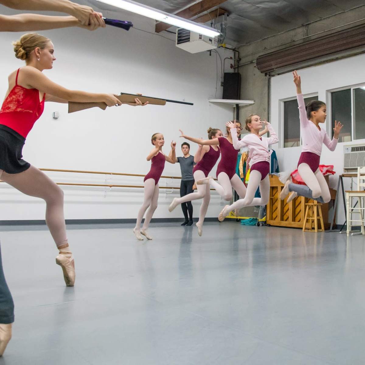 A group of ballet dancers practices in a studio, some jumping while others hold positions, with an instructor observing.