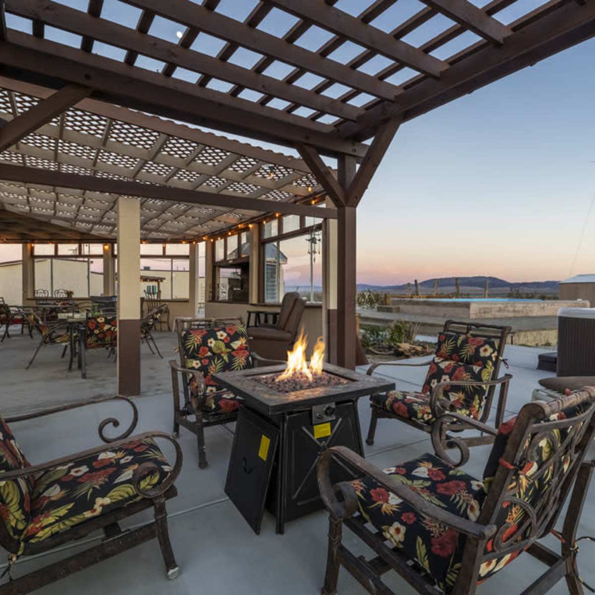 A spacious rooftop area with wooden pergolas, seating around a fire pit, and mountain views in the background during sunset.
