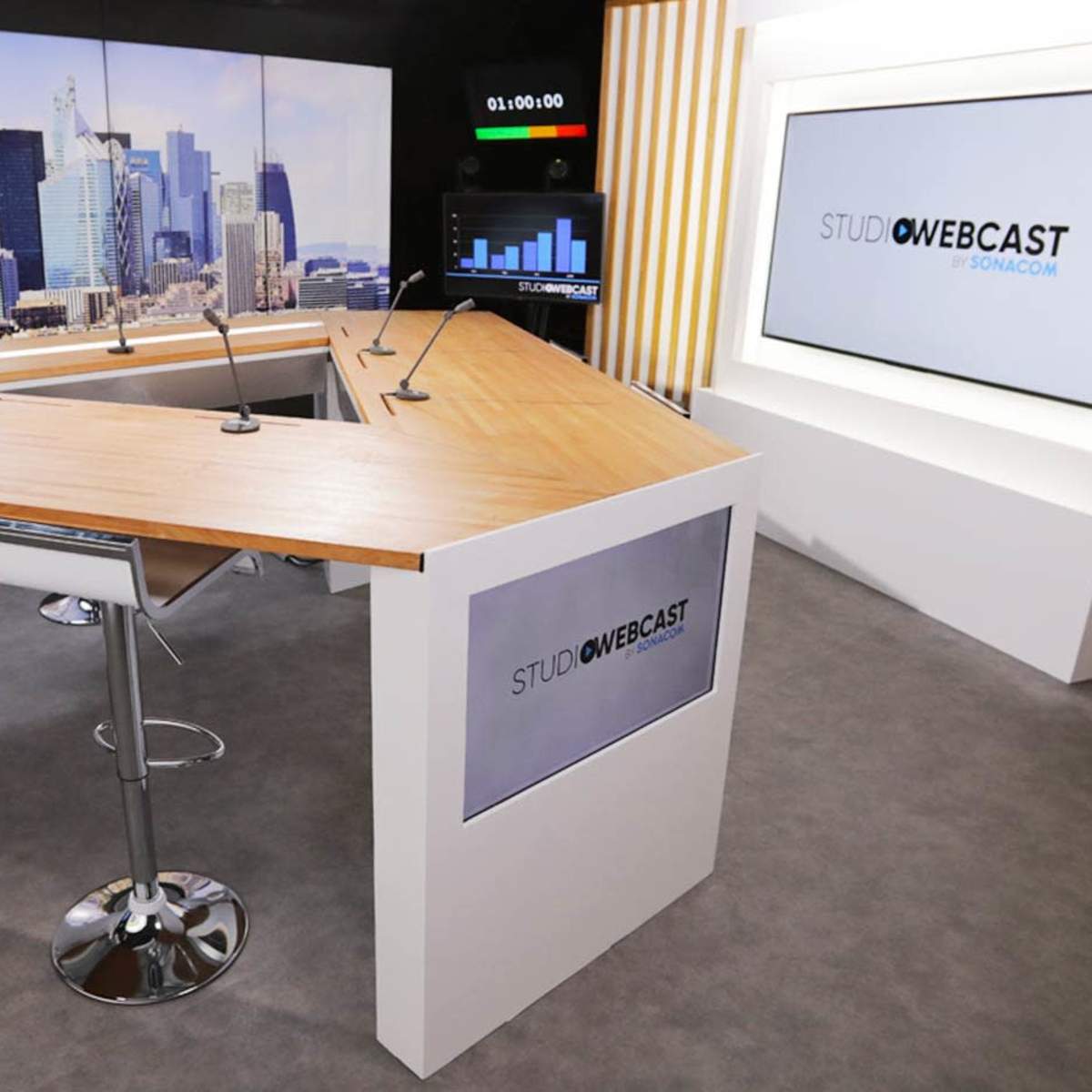 A modern broadcast studio with a segmented desk and tall bar stools, set against a backdrop of a city skyline.