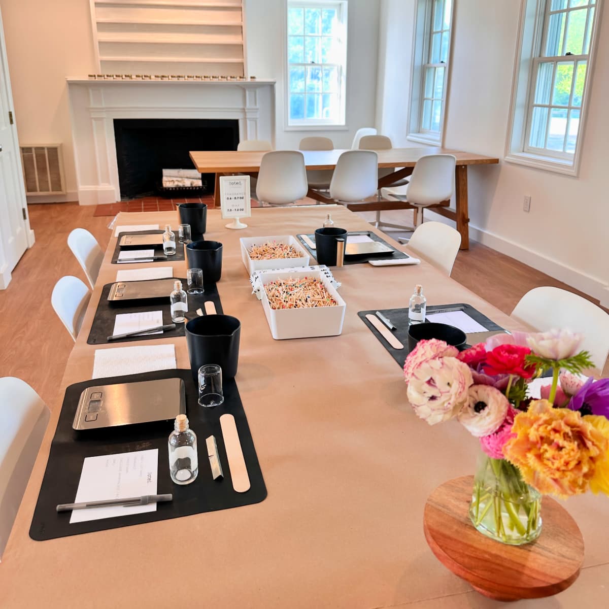 A meeting room features a long table set with water bottles, notepads, and a decorative floral arrangement, complemented by a fireplace and large windows.