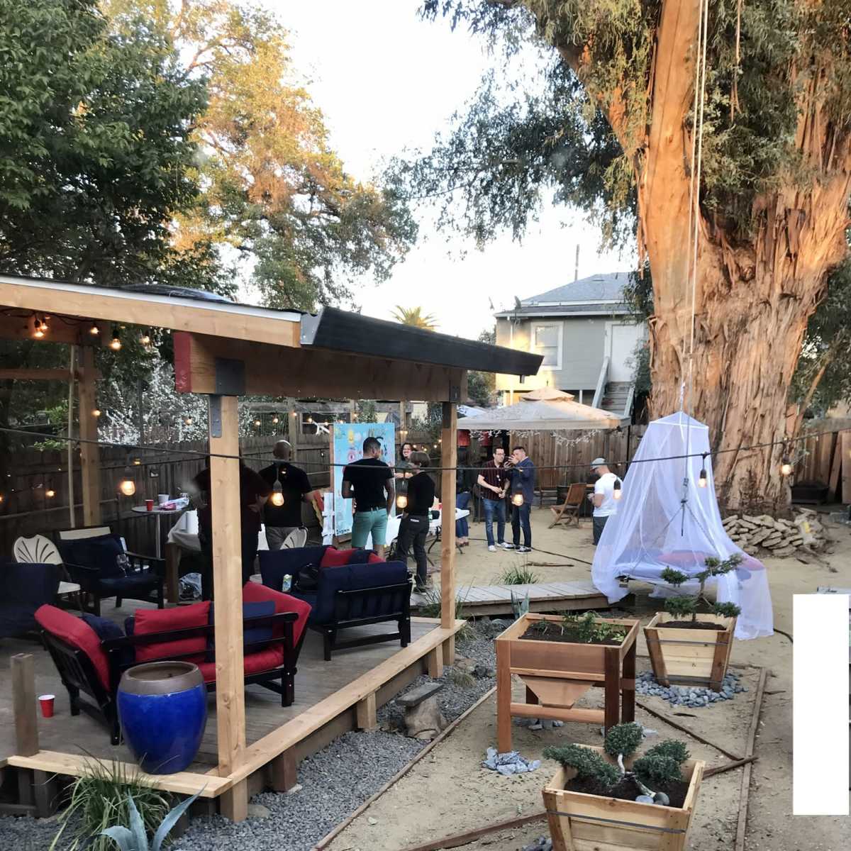 A backyard with a wooden patio area, several people gathered, and garden beds with small plants.