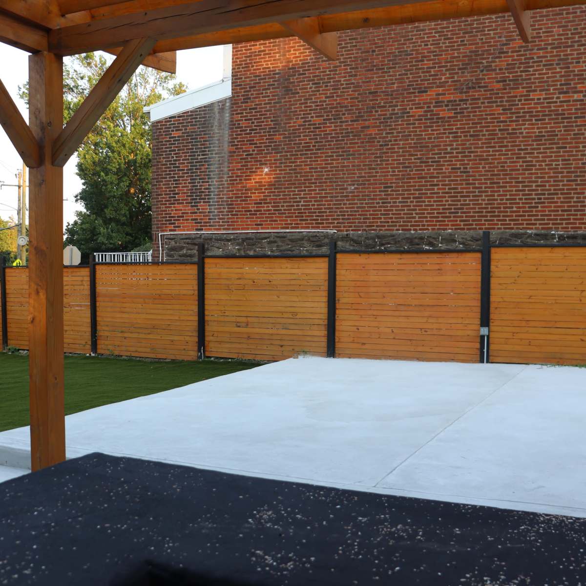 A patio area with a wooden pergola, concrete flooring, and a grassy section enclosed by wooden fencing and a brick building in the background.