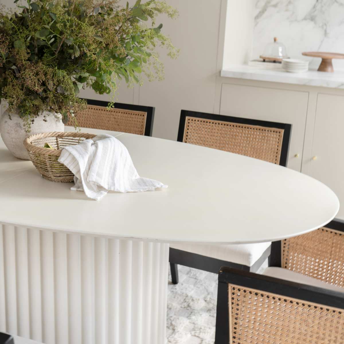 A minimalistic dining area features an oval table with a striped cloth and a basket on it, surrounded by four cane-back chairs and a large vase filled with greenery.