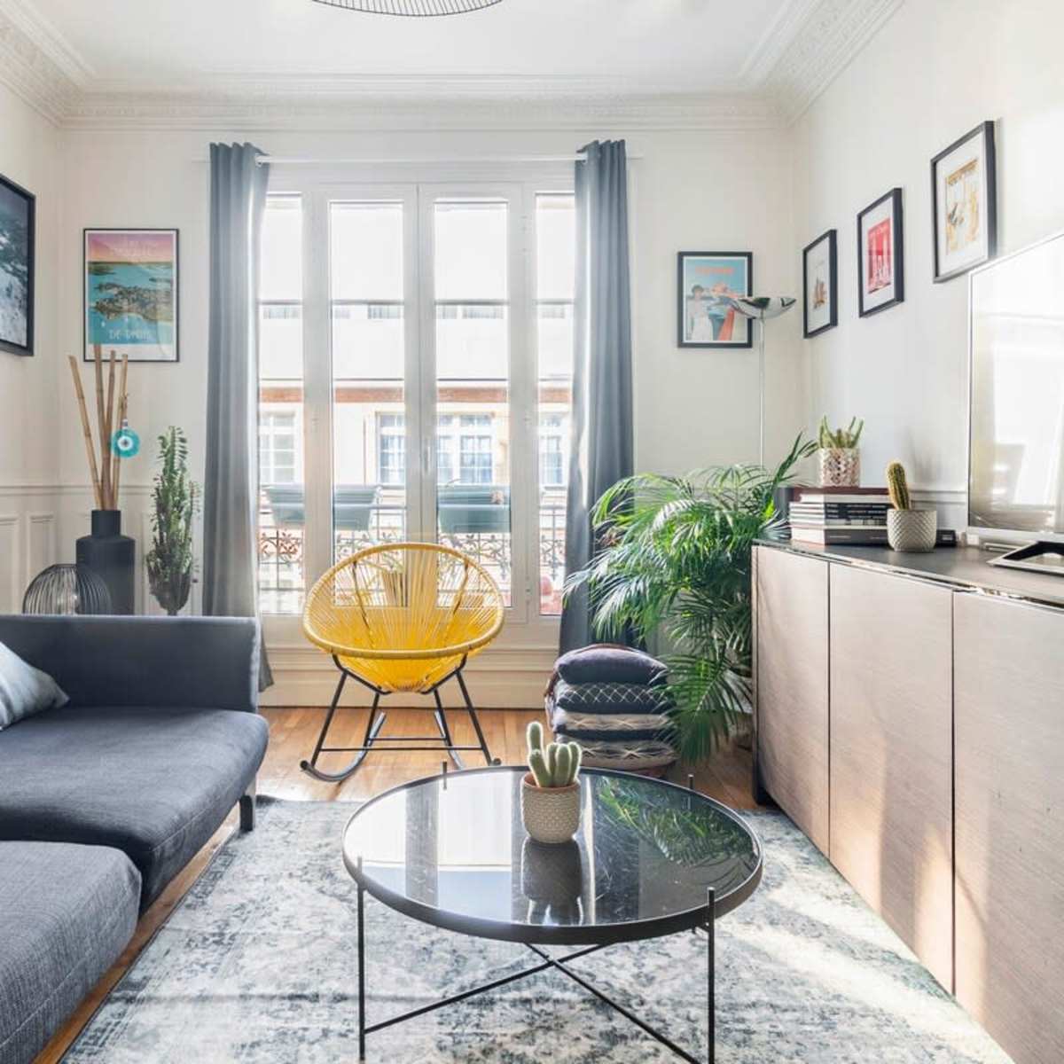 A modern living room features a dark sofa, a yellow chair, a round black coffee table, and various plants, with framed artwork hanging on the walls.