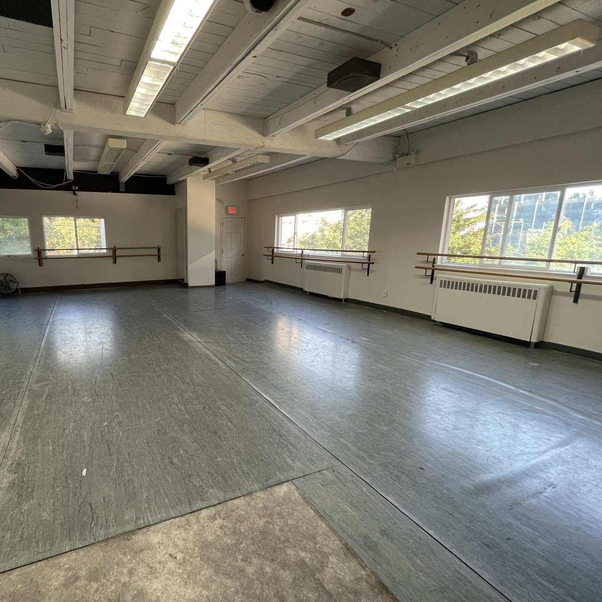 A spacious dance studio with wooden ballet barres along the walls, large windows allowing natural light, and gray flooring.