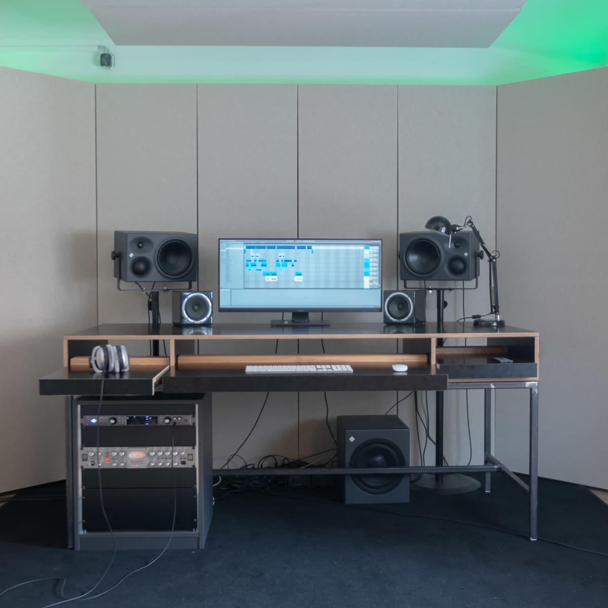 A modern music production studio features a desk with a computer monitor, audio speakers, and sound equipment against soundproofed walls.