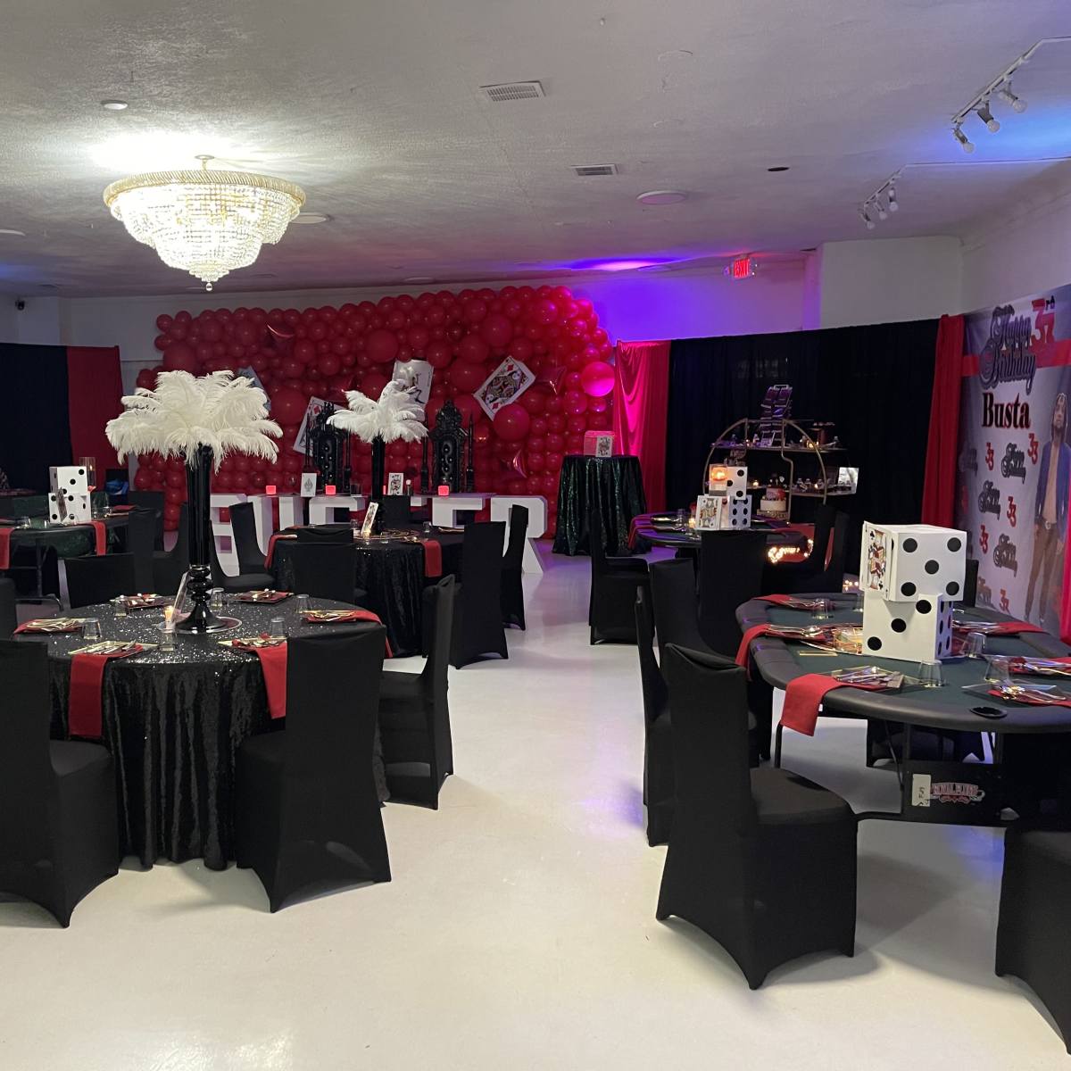 The room is set up for an event, featuring black and red tables adorned with decorations, large dice, and feather centerpieces, surrounded by balloon arches.