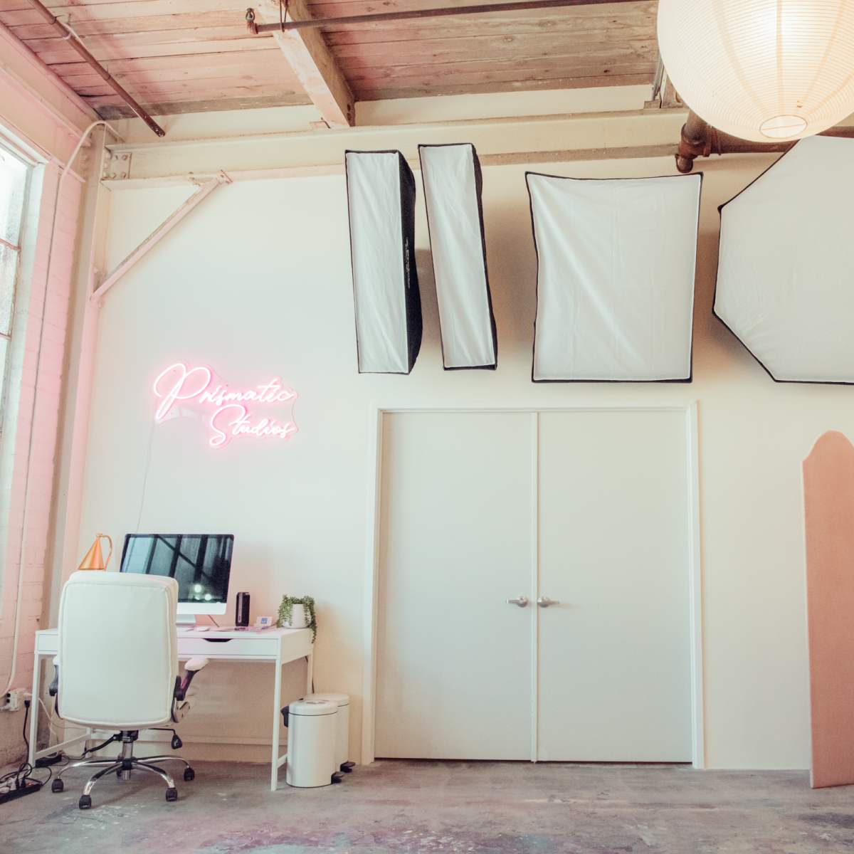 A bright photography studio featuring a workspace with a desk and computer, along with large softboxes and a decorative pink partition on the wall.