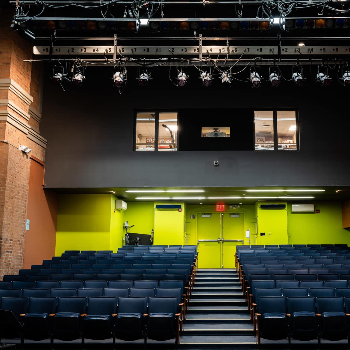 A theater auditorium with rows of blue seats facing a stage, overhead lighting, and green and brown walls.