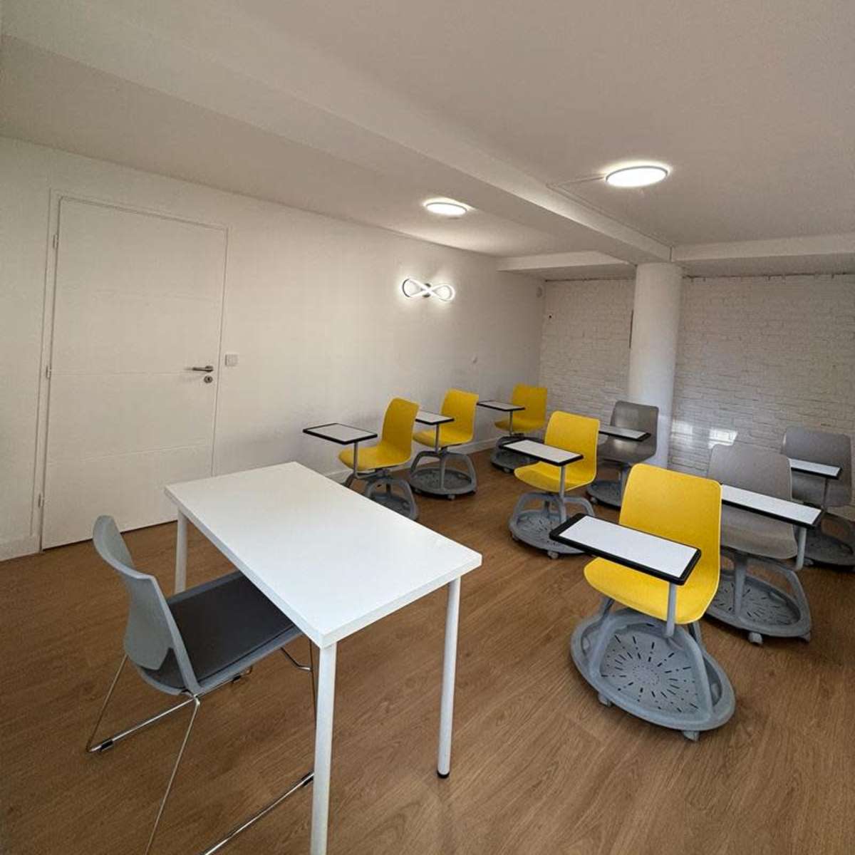 A brightly lit classroom with several yellow and gray chairs positioned around white tables.