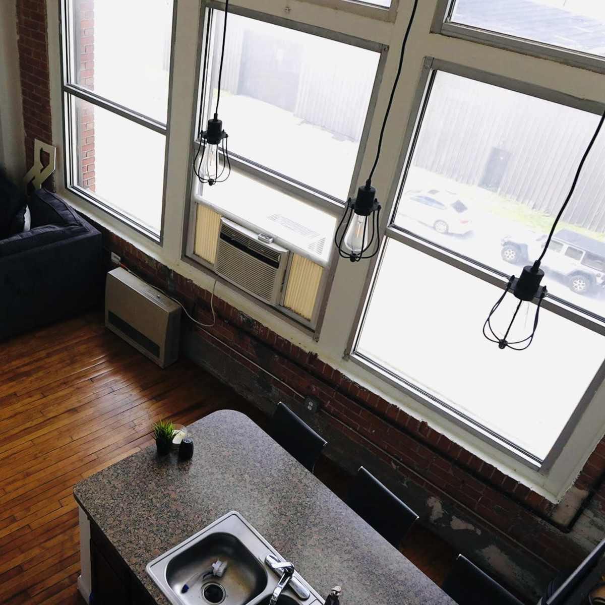 A loft-style living space with large windows, a dark sofa, a kitchen island with a sink, and a view of parked cars outside.