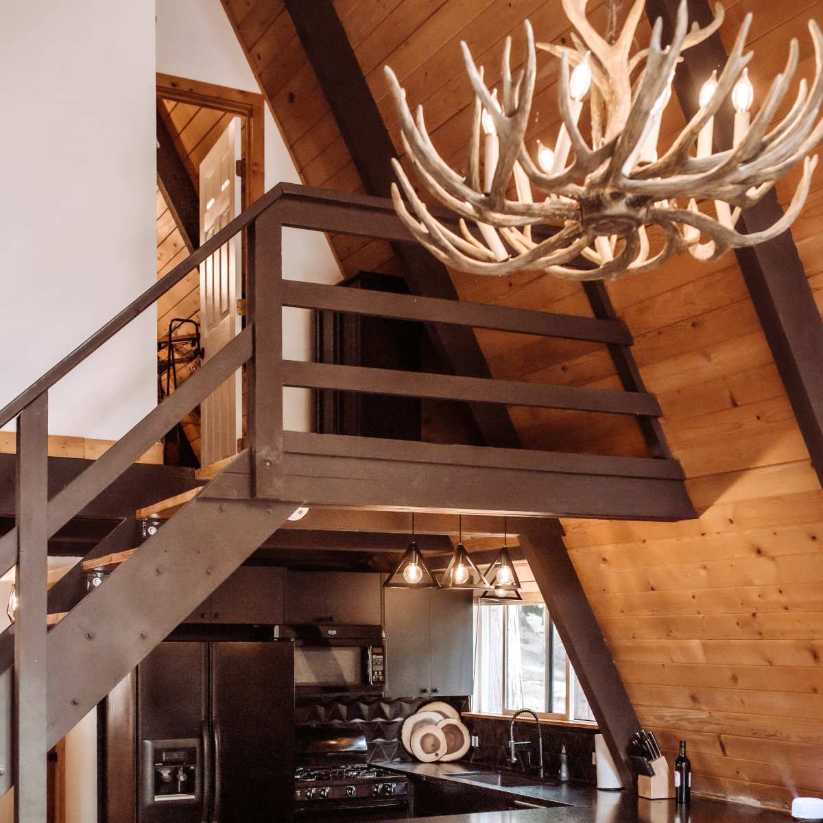 A modern cabin interior featuring an open kitchen with a black countertop, a dining area with black bar stools, and a loft area illuminated by a large antler chandelier.