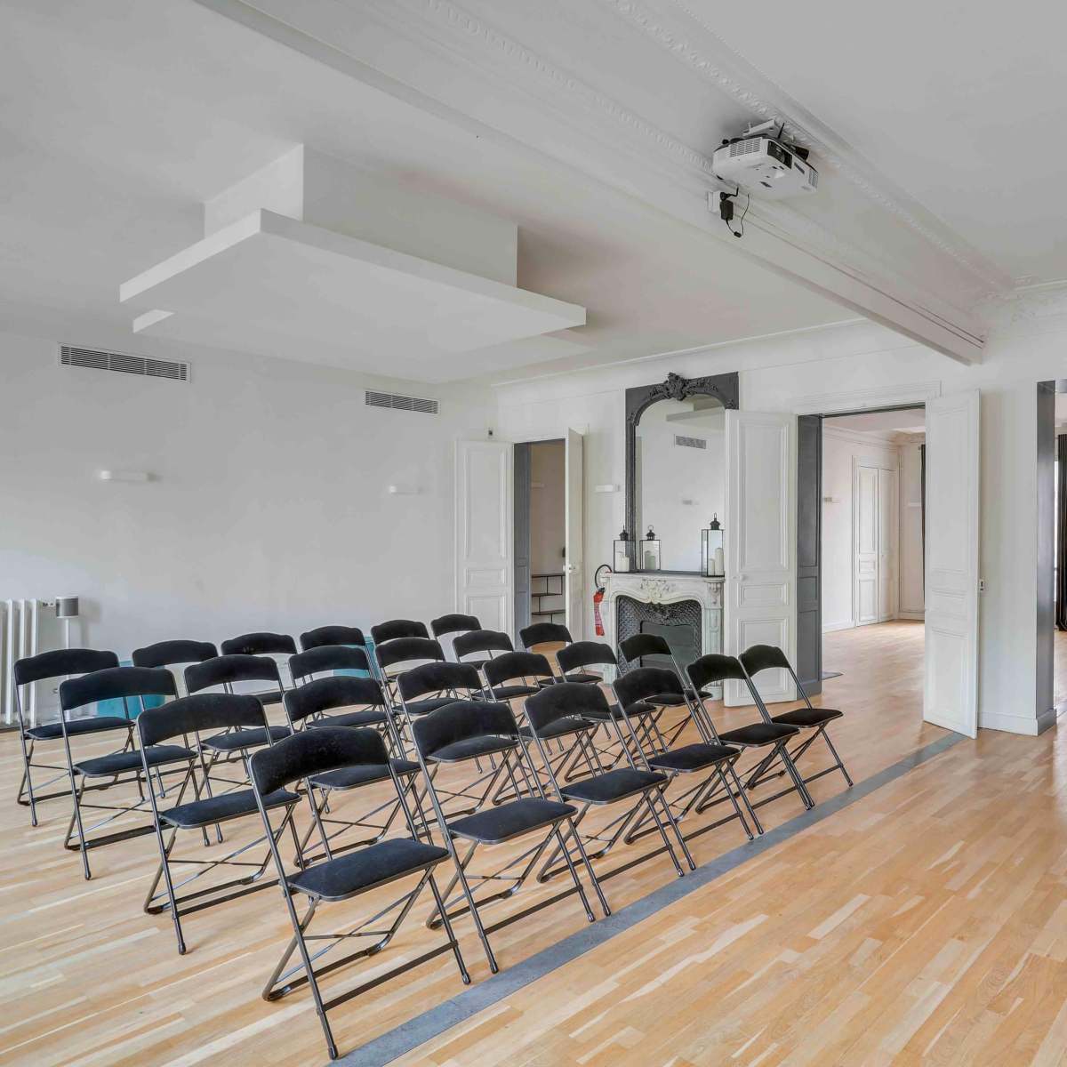A spacious room arranged with rows of black folding chairs, a small seating area in the corner, and large windows allowing natural light.